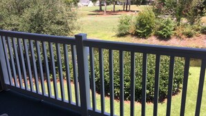 Une vue depuis un balcon doté d’une balustrade blanche, donnant sur une pelouse bien entretenue, agrémentée d’arbres et d’arbustes, et sur un bâtiment blanc situé au loin.