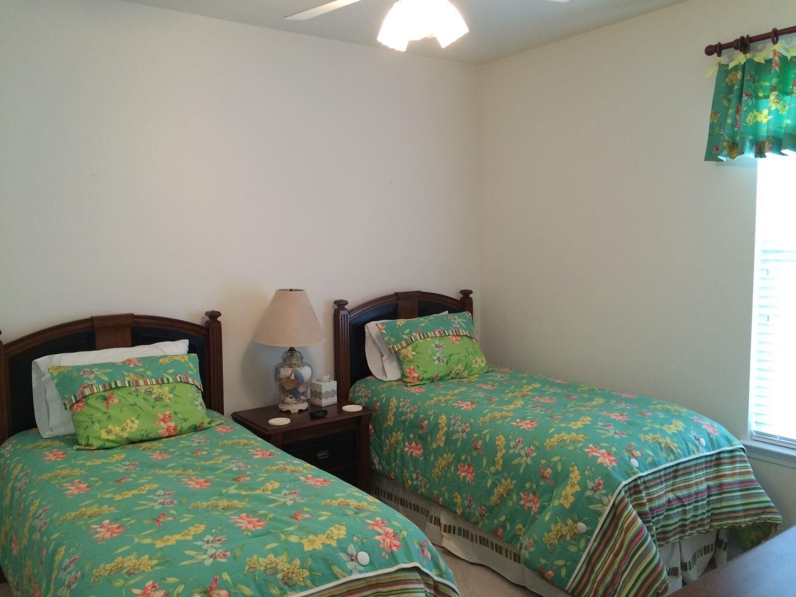 A bedroom with two single beds, each with floral bedding, a bedside table with a lamp, and a window with curtains.