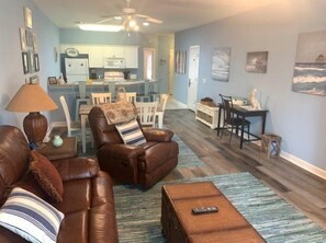 A living room with a brown leather sofa, a coffee table, and a lamp. There is a dining area with a table and chairs, and a kitchen in the background.