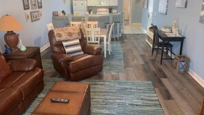 A living room with a brown leather sofa, a brown armchair with a patterned throw, a small table with a remote control, and a striped rug. There is a kitchen in the background with white cabinets and a dining area with a table and chairs. - 3709 Champions (Sunset Beach)