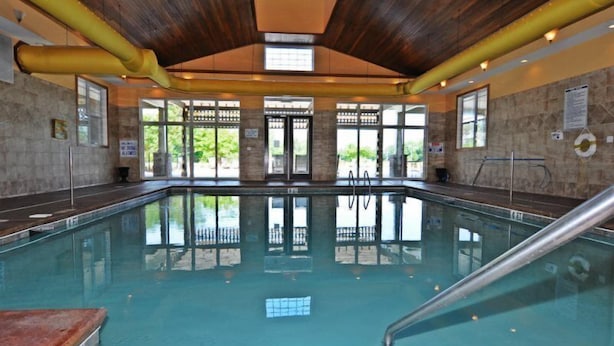 Une piscine intérieure aux parois en verre transparent, avec des poutres en bois au plafond et une vue sur l’extérieur grâce à de grandes fenêtres.