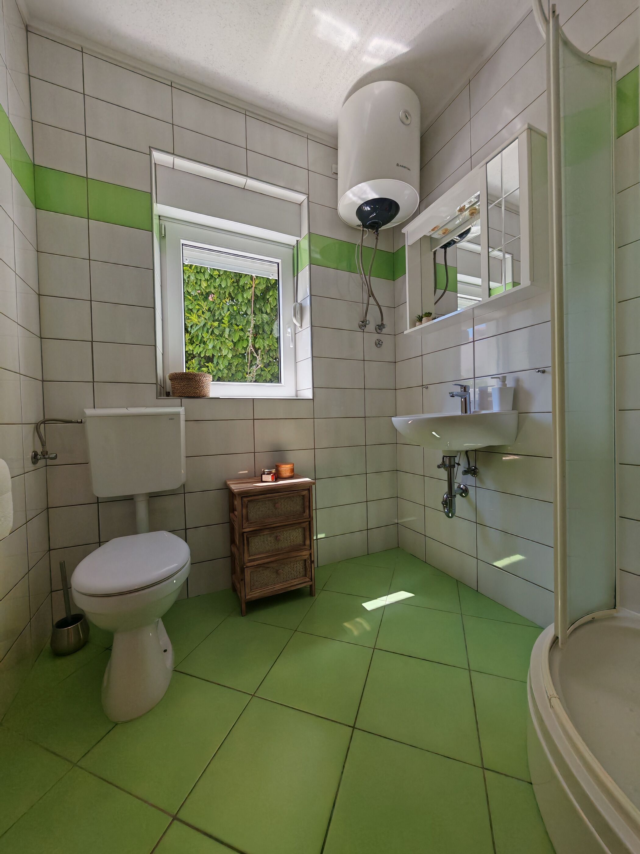 A bathroom with a toilet, a small wooden cabinet, a window with a view of greenery, a mirror, and a shower area.