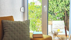 Een kamer met een bank, een kussen, een boek, een vaas met gedroogde planten en uitzicht op groen buiten.