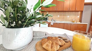 Een keuken met houten kastjes, een wit aanrecht, een bloempot, een houten plank met croissants en een glas sinaasappelsap.