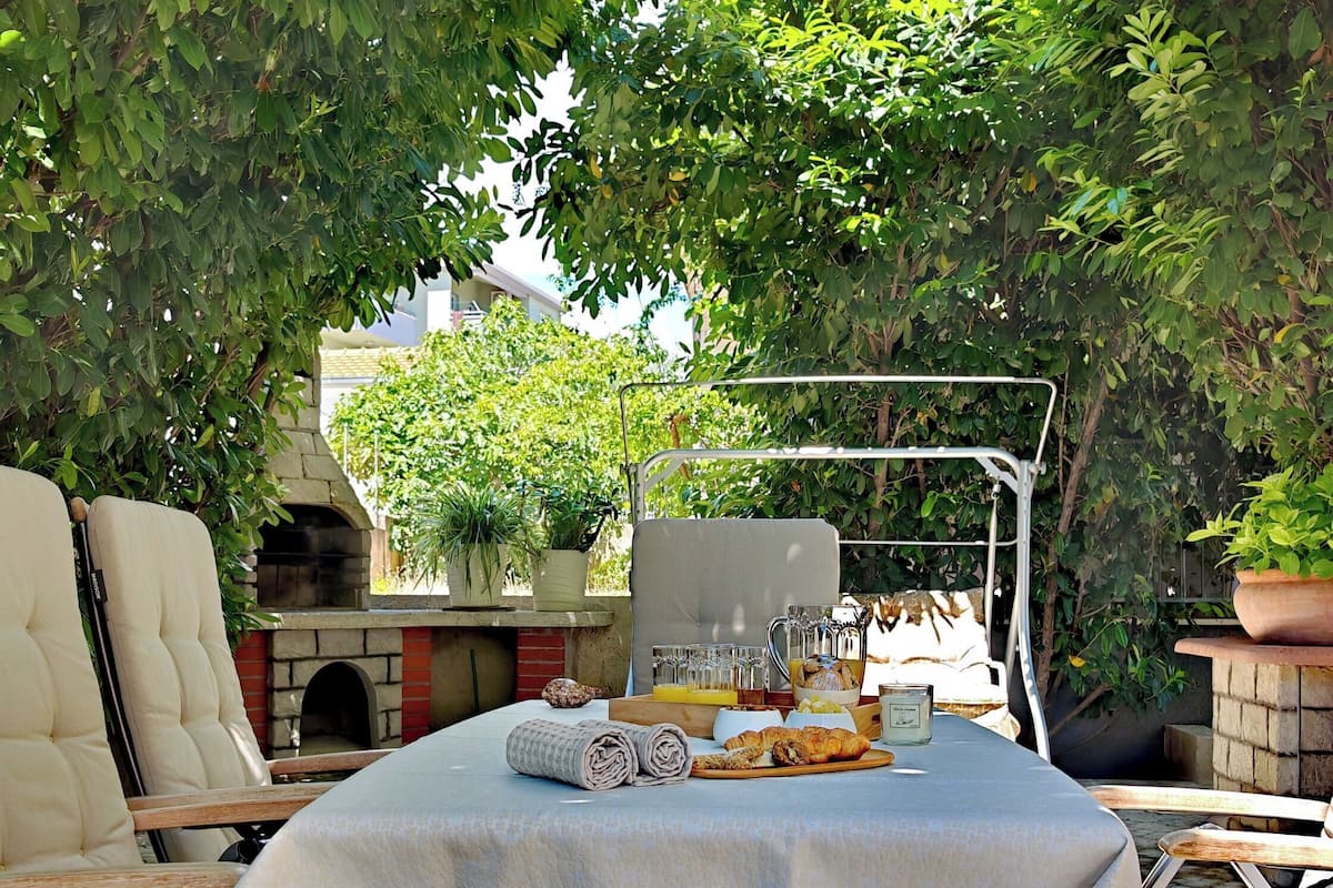 Une terrasse extérieure avec une table dressée pour le repas, entourée de chaises, et un barbecue à l’arrière-plan.