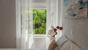 Een slaapkamer met een bed, een raam met dunne gordijnen, een vaas met bloemen en een schilderij aan de muur.