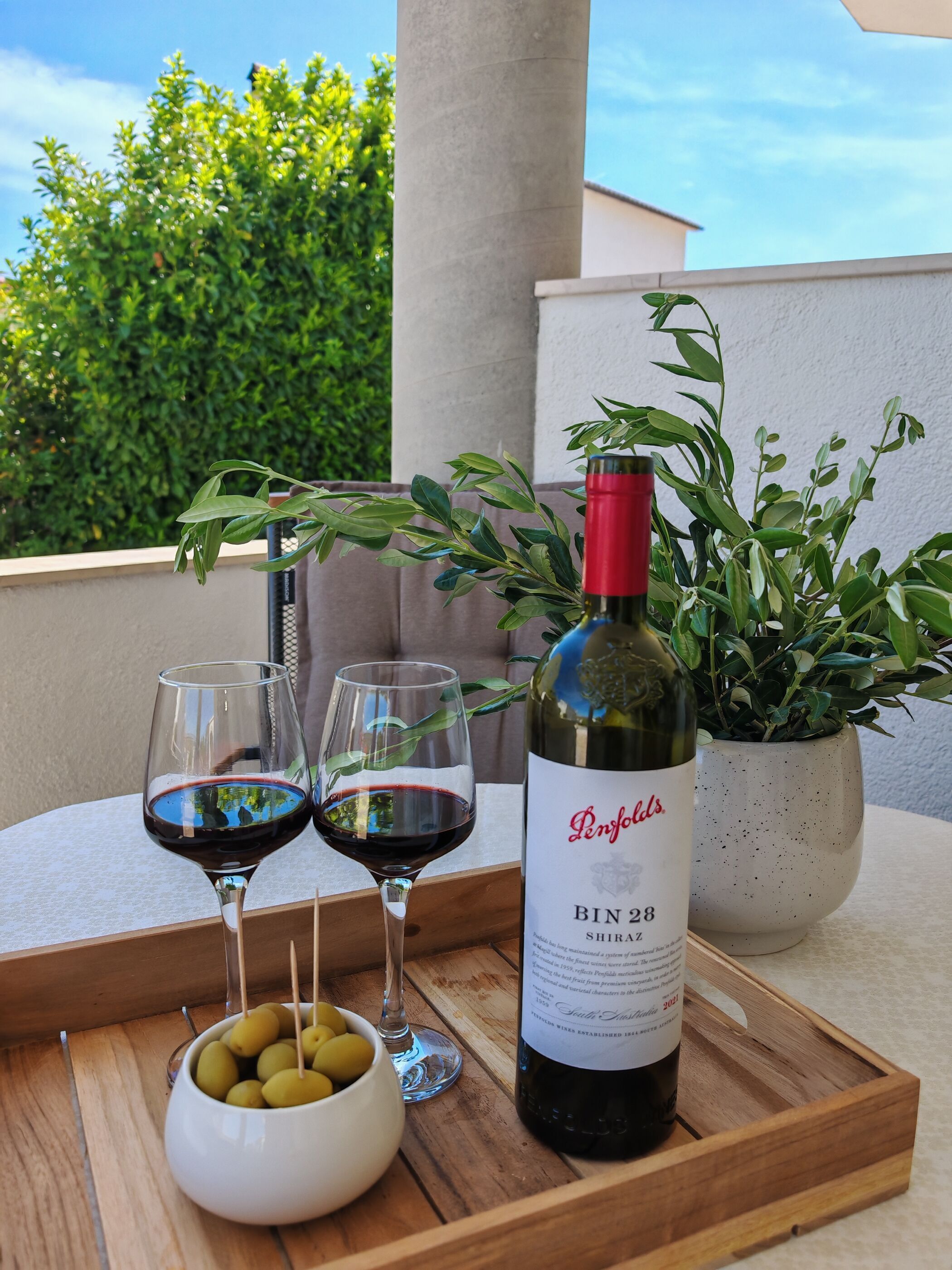 A wooden tray with two wine glasses, a bottle of wine, and a bowl of olives on a balcony.