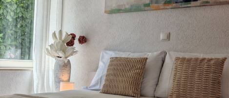 A neatly made bed with a folded towel, a painting of a lighthouse, and a vase with flowers on the wall.