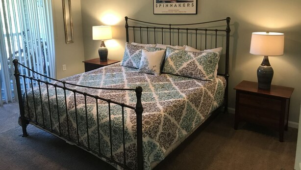 A neatly made bed with a patterned bedspread, two bedside tables with lamps, a framed picture on the wall, and a window with blinds. - 22C Sandpiper Bay (Sunset Beach)