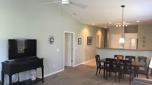 A living room with a TV, a ceiling fan, a dining area with chairs and a table, and a kitchen area with a counter and cabinets. - 22C Sandpiper Bay (Sunset Beach)