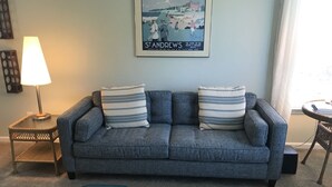 A living room with a grey sofa, two striped pillows, and two matching ottomans. A framed picture of St. Andrews is on the wall. - 22C Sandpiper Bay (Sunset Beach)