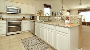 A kitchen with white cabinetry, a double oven, a microwave, a dishwasher, granite countertops, a patterned rug, and a ceiling fan. - 2407 Commons II (Calabash)