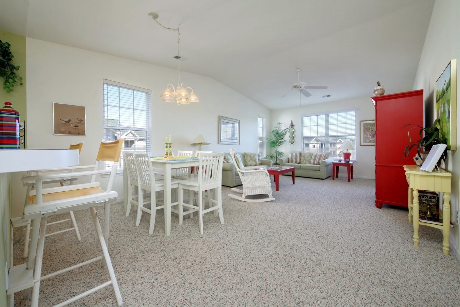 A spacious living room with a high ceiling, white furniture, a red refrigerator, and a dining area with a table and chairs.