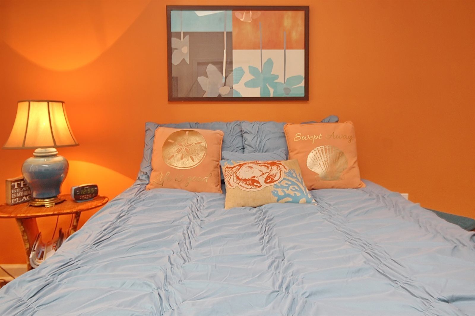 Quarto com cama, almofadas, mesa de cabeceira, luminária e uma obra de arte na parede.