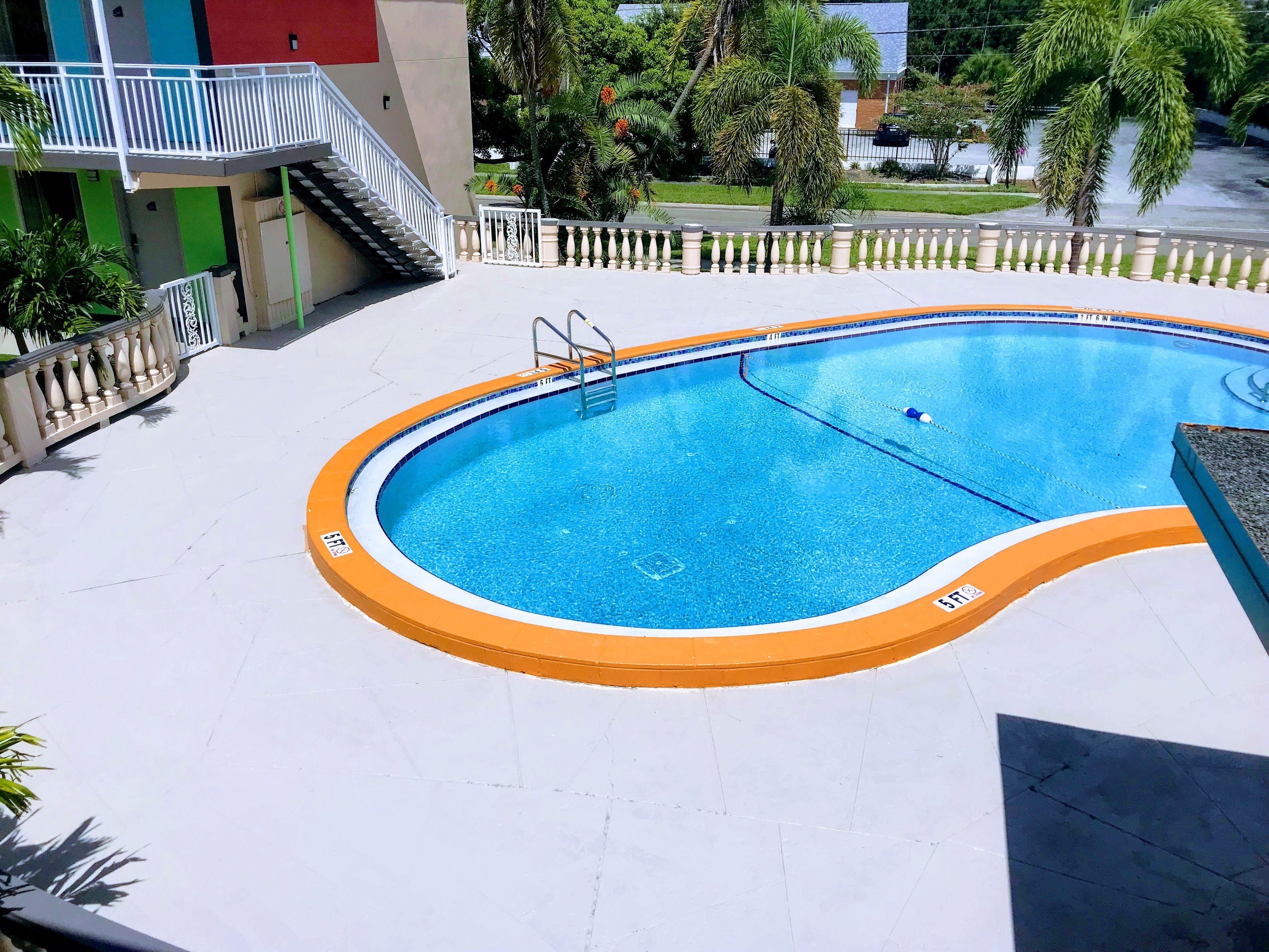 Ein Swimmingpool mit weiß-orangefarbener Umrandung, umgeben von einer weißen, gefliesten Sonnenterrasse und einem weißen Geländer.