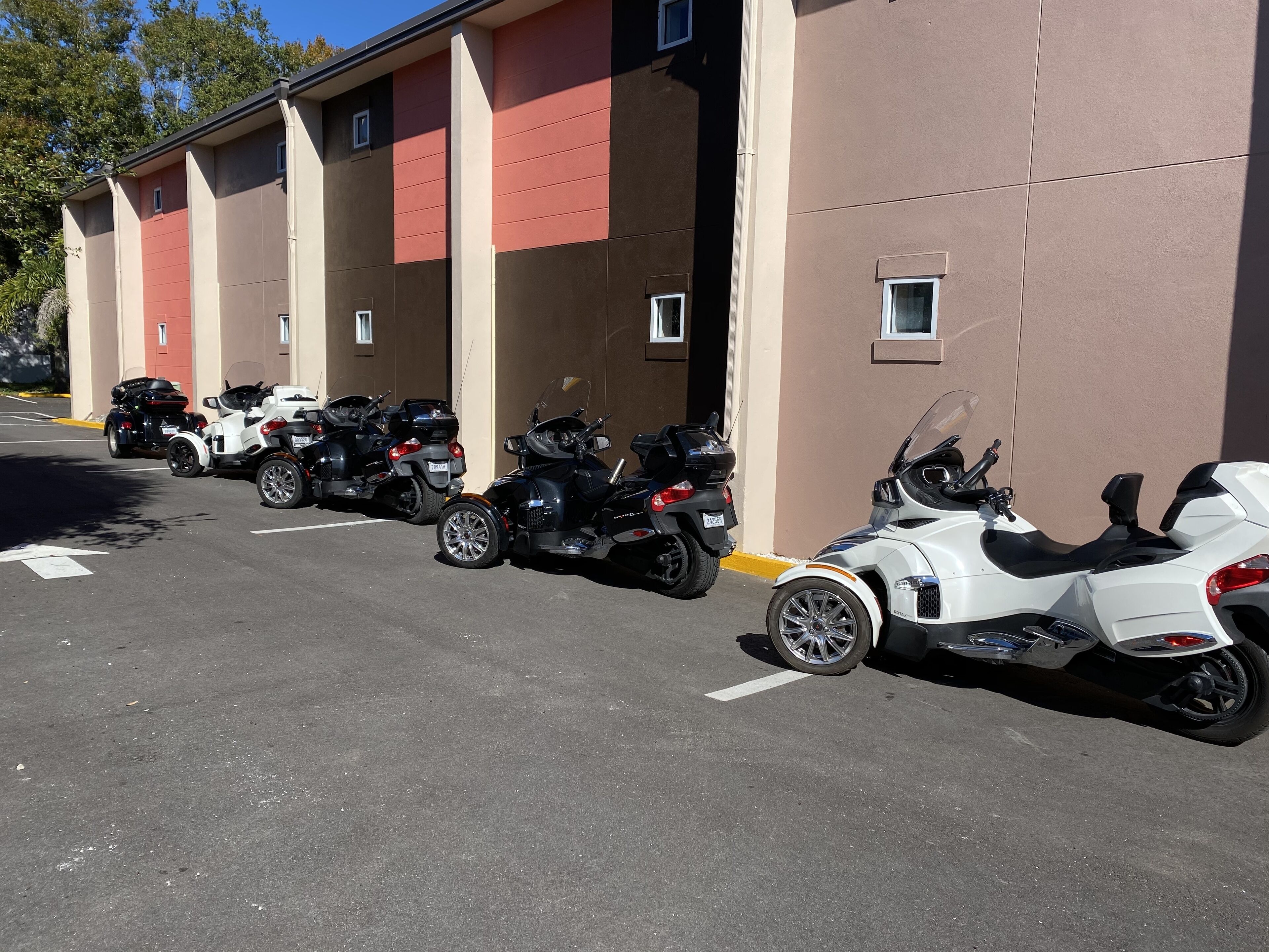 A row of parked motorcycles in a parking lot adjacent to a building with a beige and red facade.