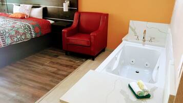 A hotel room with a bed, a red armchair, a white bathtub, and a small table with a towel and sponge.