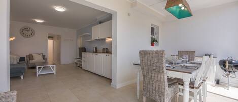 A spacious living area with a dining table, wicker chairs, a sofa, and a kitchen in the background.