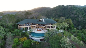 Une grande maison à deux étages, dotée d’une piscine, entourée d’une végétation luxuriante et de collines.