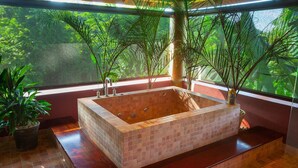 Un bain à remous rectangulaire, dont l’intérieur est revêtu de carreaux, entouré de plantes en pot, et offrant une vue sur une végétation luxuriante à travers une fenêtre.