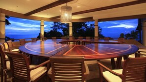 Une grande table de conférence en bois, entourée de chaises, se trouve dans un vaste espace extérieur offrant une vue sur l’océan au crépuscule.