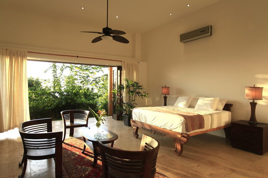 A bedroom with a bed, two bedside tables, a ceiling fan, and a view of greenery outside.