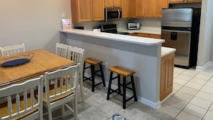 A kitchen with wooden cabinets, a stainless steel refrigerator, a microwave, and a toaster. There is a white countertop and a wooden dining table with chairs. - 1115 Crow Creek Drive (Calabash)