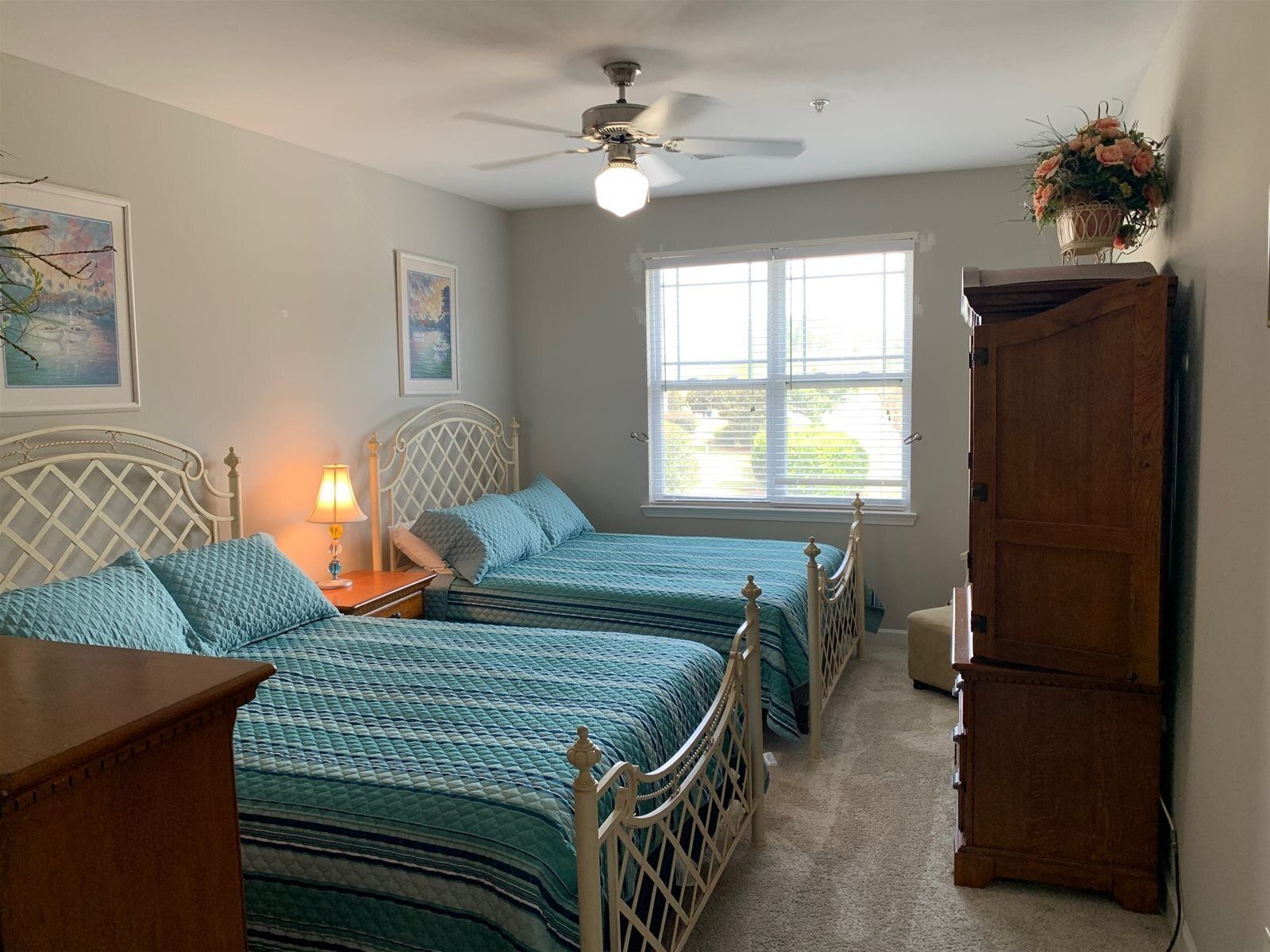 A bedroom with two beds, a ceiling fan, a window with blinds, and a wooden cabinet.