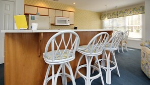 Een keuken met een witte koelkast, magnetron en houten lambrisering. Er staan vier witte stoelen met bloemenkussentjes rond een klein eiland.