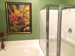 A bathroom with a glass-enclosed shower, a white bathtub, and a framed tropical painting on the wall.