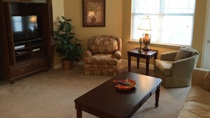 Una sala de estar con un mueble para televisor, dos sillones, una mesa de centro, una lámpara y una ventana con persianas.