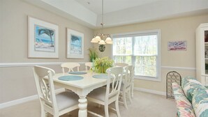 A dining area with a white table, four chairs, and a chandelier. There are framed pictures on the wall and a window with blinds. - 805 Colony II (Sunset Beach)