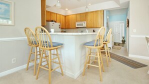 A kitchen with wooden cabinets, a white refrigerator, a microwave, and two bar stools with blue cushions. - 805 Colony II (Sunset Beach)