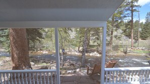 A covered outdoor area with a wooden deck, two wooden chairs, and a view of a rocky stream surrounded by trees.
