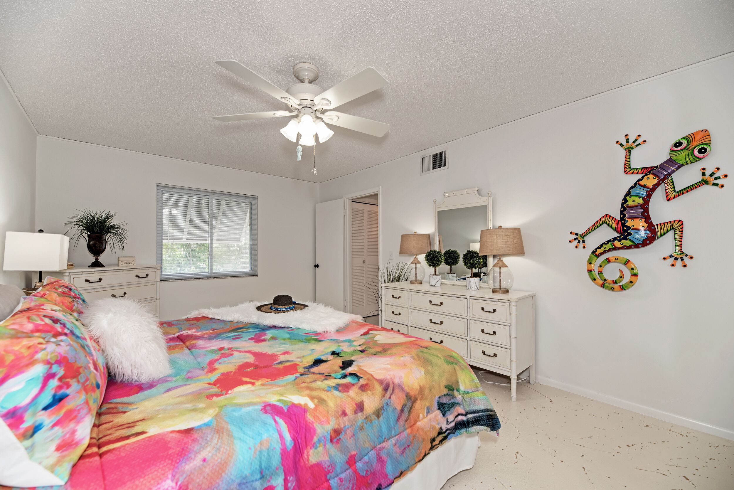 A bedroom with a colorful bedspread, a ceiling fan, a mirror, and a decorative lizard on the wall.