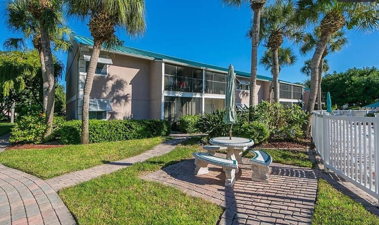A building with a modern design, featuring a flat roof and large windows. There is a paved pathway leading to a seating area with a table and chairs.