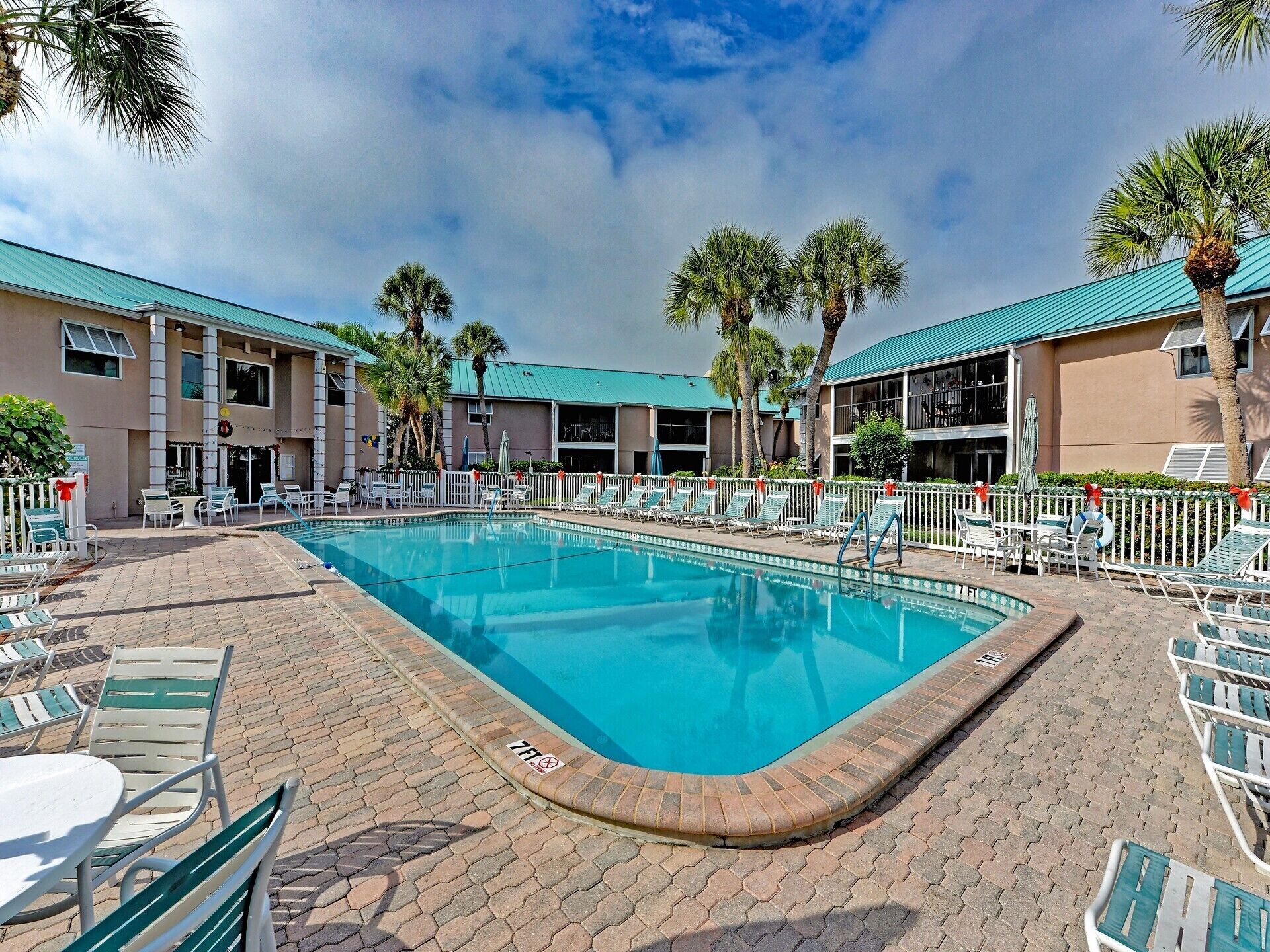 A swimming pool with lounge chairs and palm trees, surrounded by a building with a green roof.