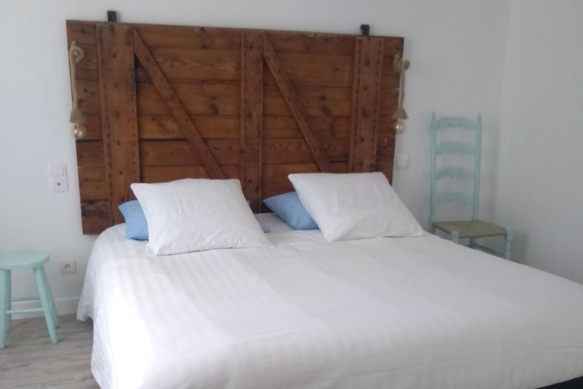 A bed with a wooden headboard, a small stool, and a chair in a room with white walls and wooden flooring.