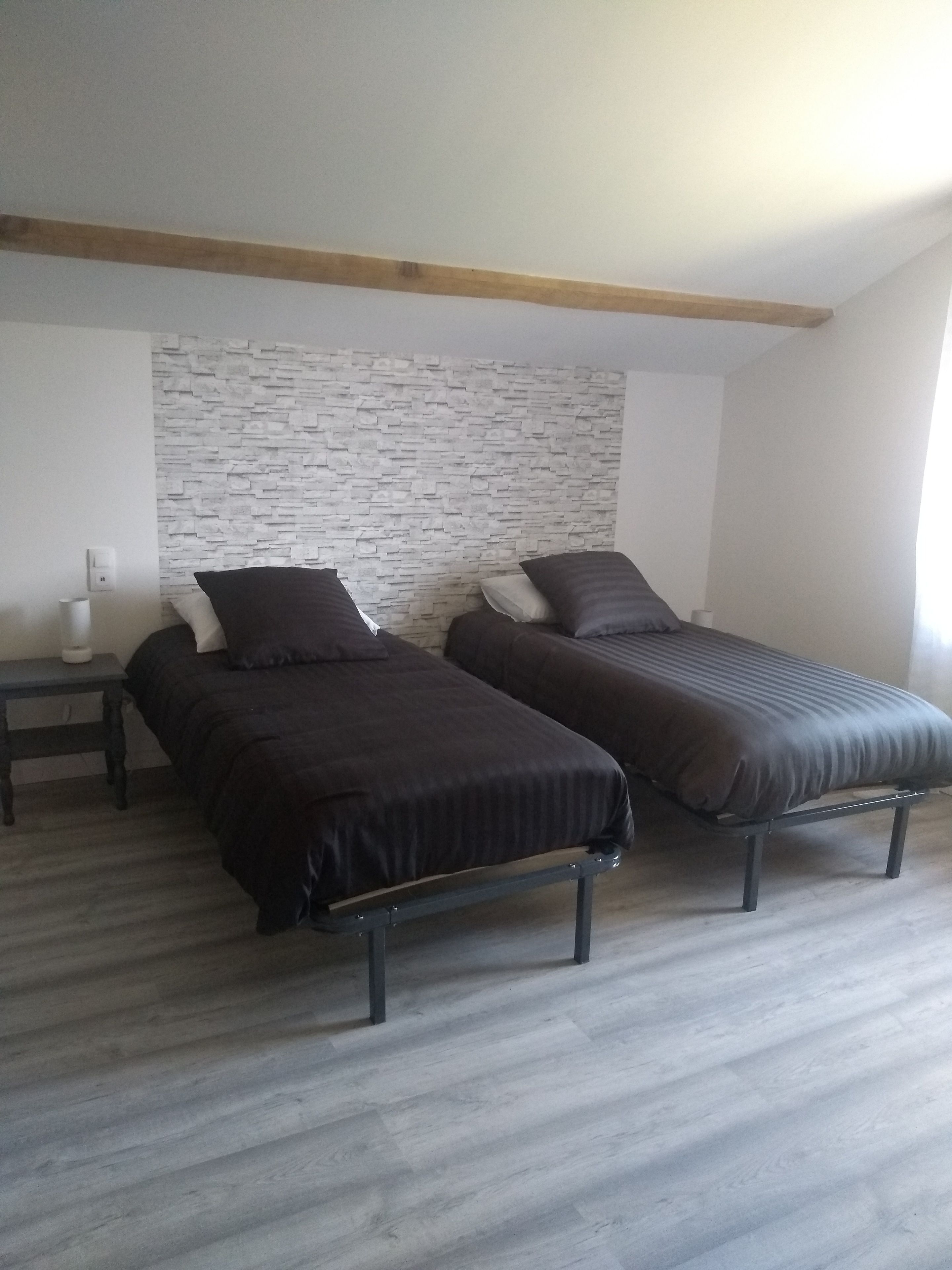 Una habitación con dos camas individuales, una mesita de noche con lámpara, un interruptor de luz en la pared y techo con vigas de madera.