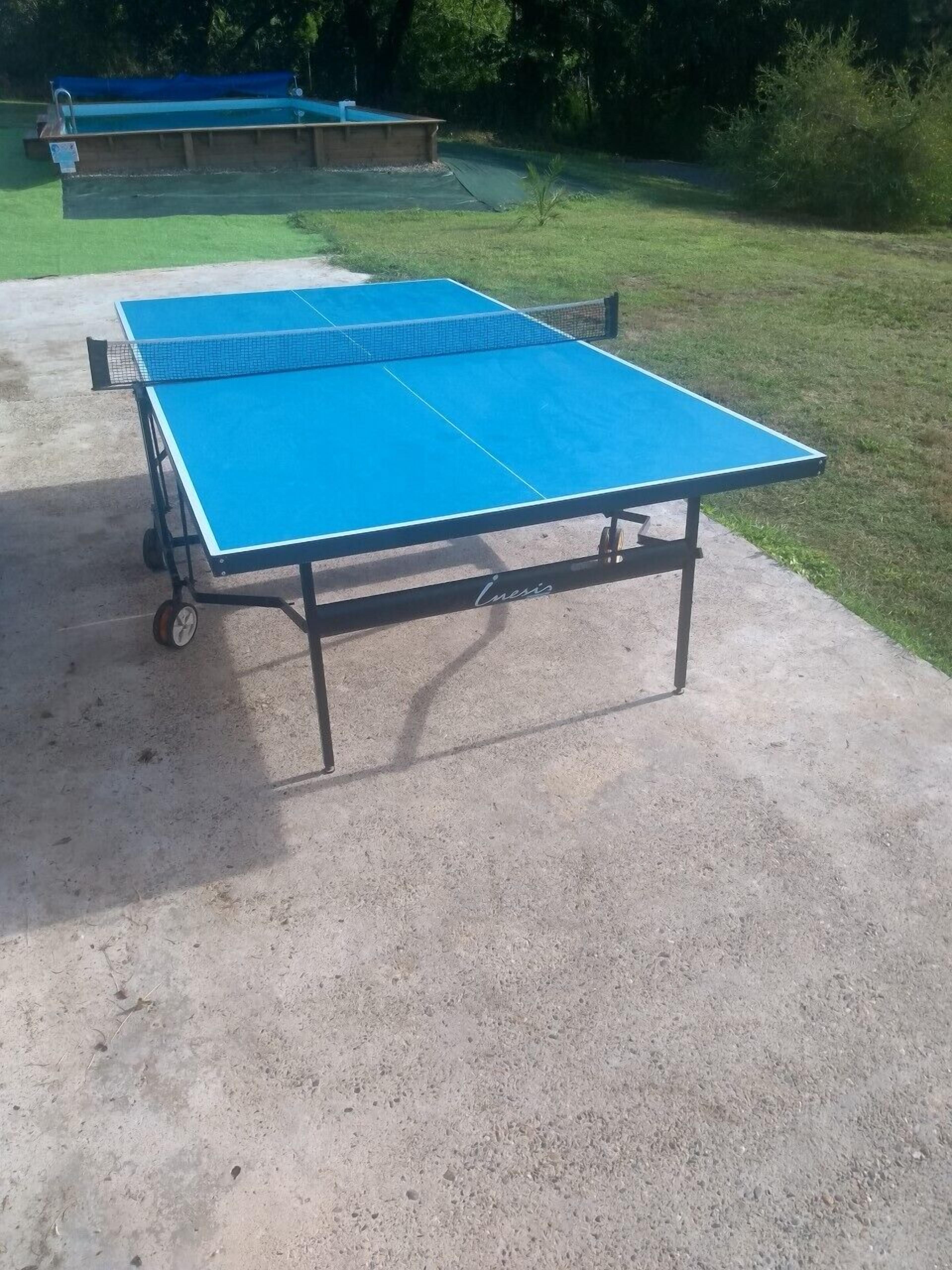 Uma mesa de pingue-pongue azul com rodinhas, colocada sobre uma superfície de concreto em área externa.