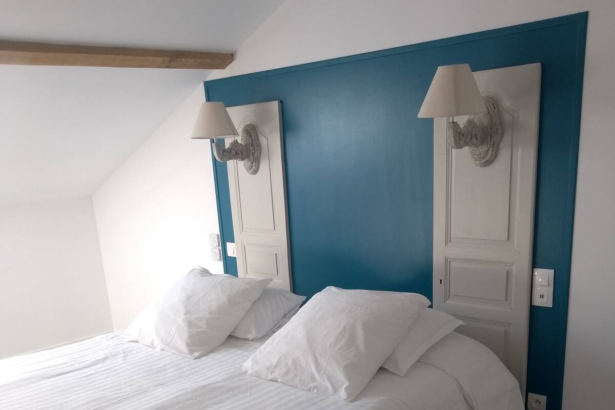 A neatly made bed with white linens in a room with a blue accent wall and a wooden ceiling beam.