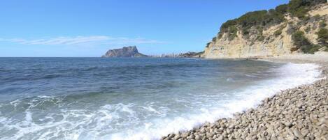Une plage de galets bordée d’une eau d’un bleu limpide, une falaise rocheuse et une île lointaine.