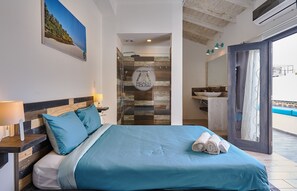 A bedroom with a bed, a nightstand, a bathroom with a sink and mirror, and a view of the beach.