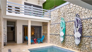A poolside area with a stone wall, two surfboards, a table with chairs, and a balcony. - Colonial Beach House (Santo Domingo)
