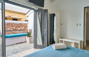 A bedroom with a bed, a bench by the window, a white console table, and a view of a brick wall outside.