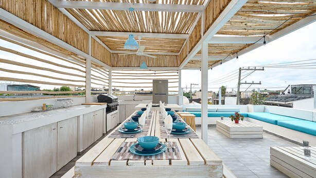An outdoor dining area with a long wooden table set for a meal, surrounded by white cabinets and a blue cushioned seating area. - Colonial Beach House (Santo Domingo)