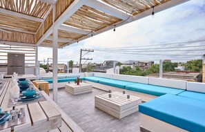 An outdoor terrace with wooden tables, blue cushions, and a view of the cityscape.