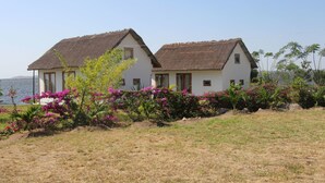 Une maison à deux étages, avec un toit de chaume, entourée d’un jardin où poussent des fleurs roses, située près d’une étendue d’eau.