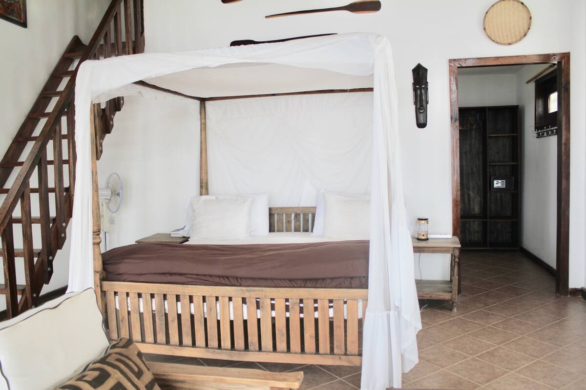 Una habitación con una cama de madera, una mesita de noche, una cortina blanca, una escalera de madera y una puerta que da a otra habitación.