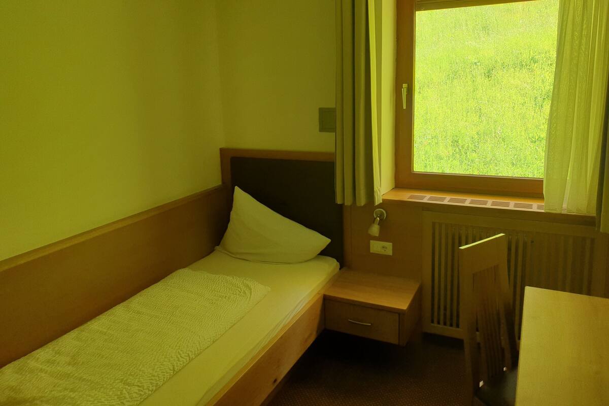 A small, single-bed room with a wooden headboard, a bedside table, and a window with curtains.